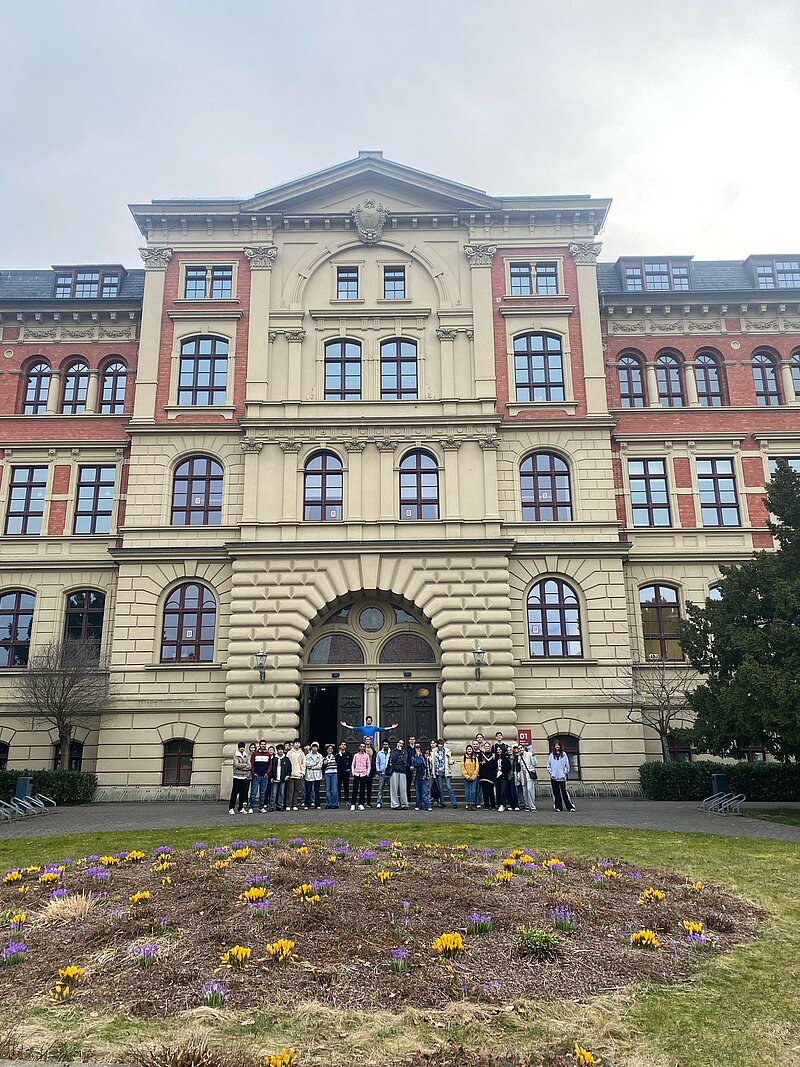 Eine Gruppe von Menschen steht vor einem großen, historischen Gebäude mit drei Eingängen und vielen Fenstern. Das Gebäude hat eine gelbe Fassade mit roten Akzenten. Im Vordergrund befindet sich ein Blumenbeet mit verschiedenen Blumen. Der Himmel ist bewölkt.