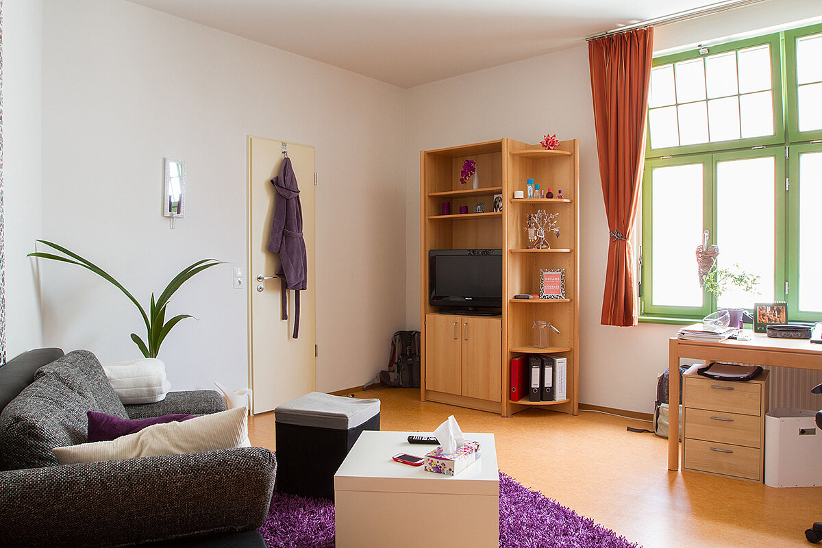 Ein gemütliches Wohnzimmer mit einem Sofa, einem Couchtisch, einem Bücherregal mit Fernseher und einem Schreibtisch. Es gibt eine Tür mit einem Garderobenhaken, eine Pflanze und bunte Vorhänge am Fenster. Der Boden ist mit einem lila Teppich bedeckt.