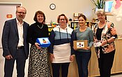 Fünf Personen stehen lächelnd nebeneinander und halten kleine Geschenke oder Urkunden in den Händen. Sie scheinen bei einer Feier oder Auszeichnung zu sein. Die Gruppe besteht aus drei Frauen und zwei Männern, die sich über ihre Erfolge freuen.