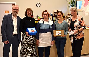 Fünf Personen stehen lächelnd nebeneinander und halten kleine Geschenke oder Urkunden in den Händen. Sie scheinen bei einer Feier oder Auszeichnung zu sein. Die Gruppe besteht aus drei Frauen und zwei Männern, die sich über ihre Erfolge freuen.