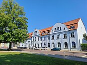 Ein großes, weißes Gebäude mit rotem Dach und vielen Fenstern. Vor dem Gebäude stehen mehrere Autos auf einem Parkplatz. Links im Bild befindet sich ein großer Baum. Der Himmel ist klar und blau, und es gibt einen grünen Rasen im Vordergrund.