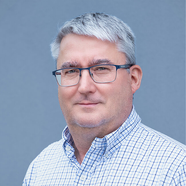 Porträt eines Mannes mit grauen Haaren, der eine Brille mit dünnem Metallgestell trägt und ein helles Hemd mit blauem Karomuster. Er lächelt leicht.