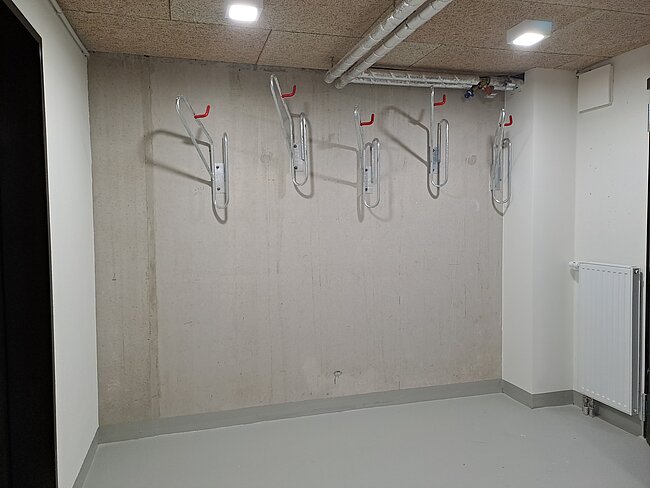 Drei silberne Fahrradhalterungen sind an einer hellgrauen Betonwand in einem Fahrradraum montiert.