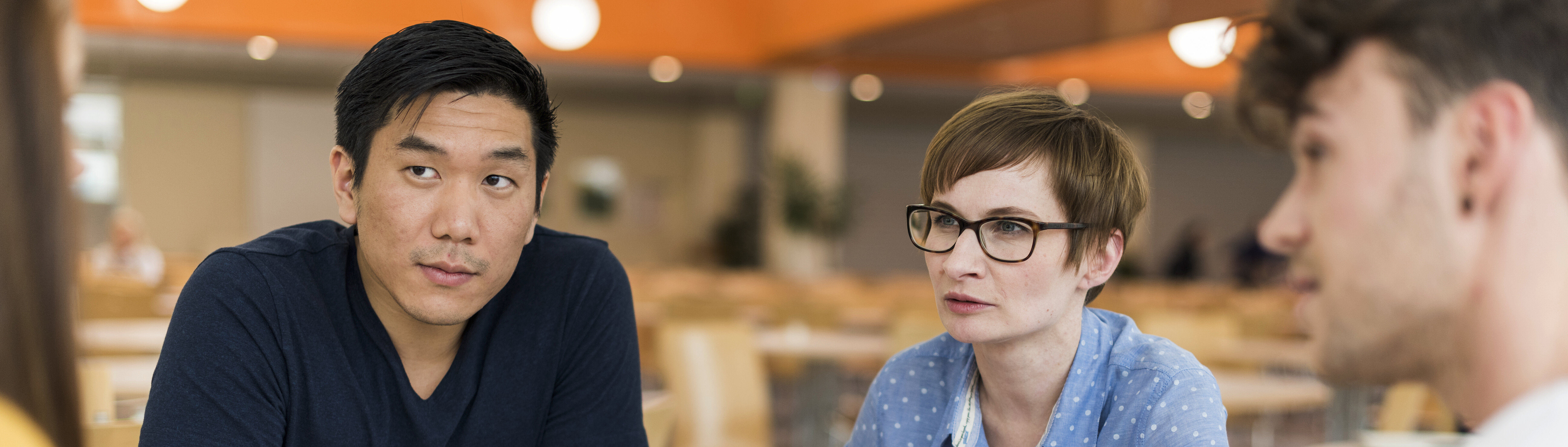 Drei Personen sitzen an einem Tisch und unterhalten sich. Eine Person zeigt auf ein Magazin, während die anderen beiden aufmerksam zuhören. Die Szene spielt in einem hellen, modernen Raum, möglicherweise einer Cafeteria oder einem Gemeinschaftsbereich.