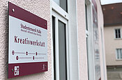 "Ein Schild an einer Wand mit der Aufschrift 'Kreativwerkstatt'. Darunter sind Kontaktdaten und Symbole für Telefon, E-Mail und eine Website. Das Schild gehört zum Studentenwerk Halle, Anstalt öffentlichen Rechts."