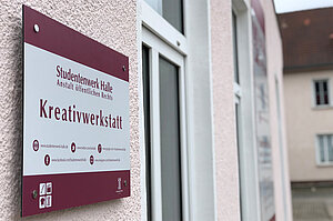 "Ein Schild an einer Wand mit der Aufschrift 'Kreativwerkstatt'. Darunter sind Kontaktdaten und Symbole für Telefon, E-Mail und eine Website. Das Schild gehört zum Studentenwerk Halle, Anstalt öffentlichen Rechts."
