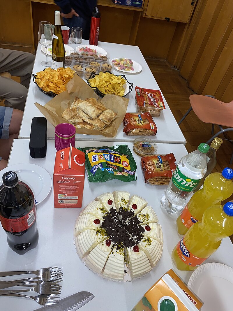Ein langer Tisch mit verschiedenen Snacks: Chips in Schälchen, Tüten mit Salzgebäck und Nüssen, eine große Torte mit Schokostückchen in der Mitte, Getränke wie Cola, Wasser und Orangensaft sowie Besteck und Servietten.