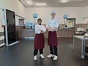 Zwei Köche in einer professionellen Küche stehend, beide in weißer Jacke, roter Schürze und weißer Mütze, mit selbstbewusstem Armkreuz vor Arbeitsinseln und Küchengeräten.