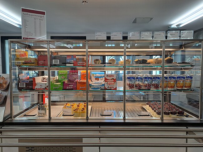 Gläser mit Süßwaren und Backwaren: Oben liegen verschiedene Kekse, Schokoladenprodukte und Snacks in Packungen, unten sind Kuchen, Torten, Muffins und Schokoladenstücke in durchsichtigen Vitrinen präsentiert.