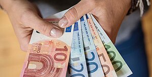 "Eine Hand hält verschiedene Euro-Scheine, darunter 10, 20, 50 und 100 Euro. Die Scheine sind fächerförmig angeordnet und zeigen die Vorderseiten mit den unterschiedlichen Designs und Farben. Die Hand hält die Scheine zwischen Daumen und Zeigefinger."