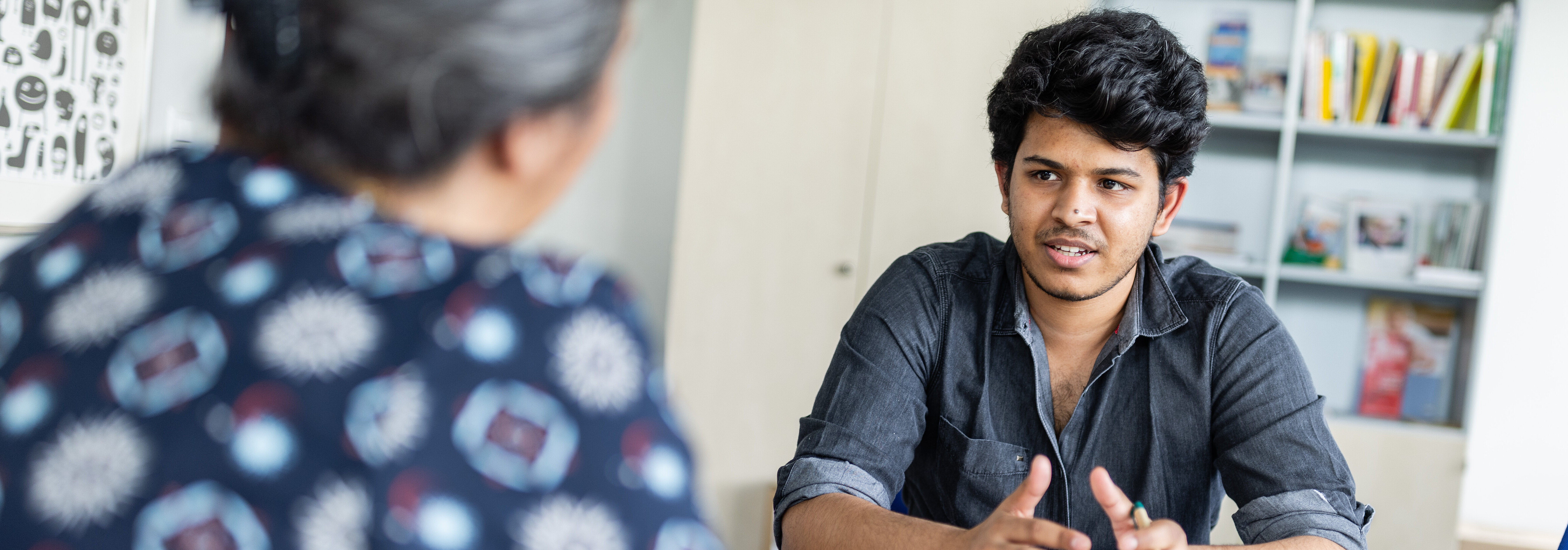 Zwei Personen sitzen sich an einem Tisch gegenüber und führen ein Gespräch. Eine Person, die teilweise zu sehen ist, trägt ein gemustertes Hemd. Die andere Person, ein junger Mann, trägt ein dunkles Hemd und gestikuliert mit den Händen. Auf dem Tisch liegen Papiere und Stifte.