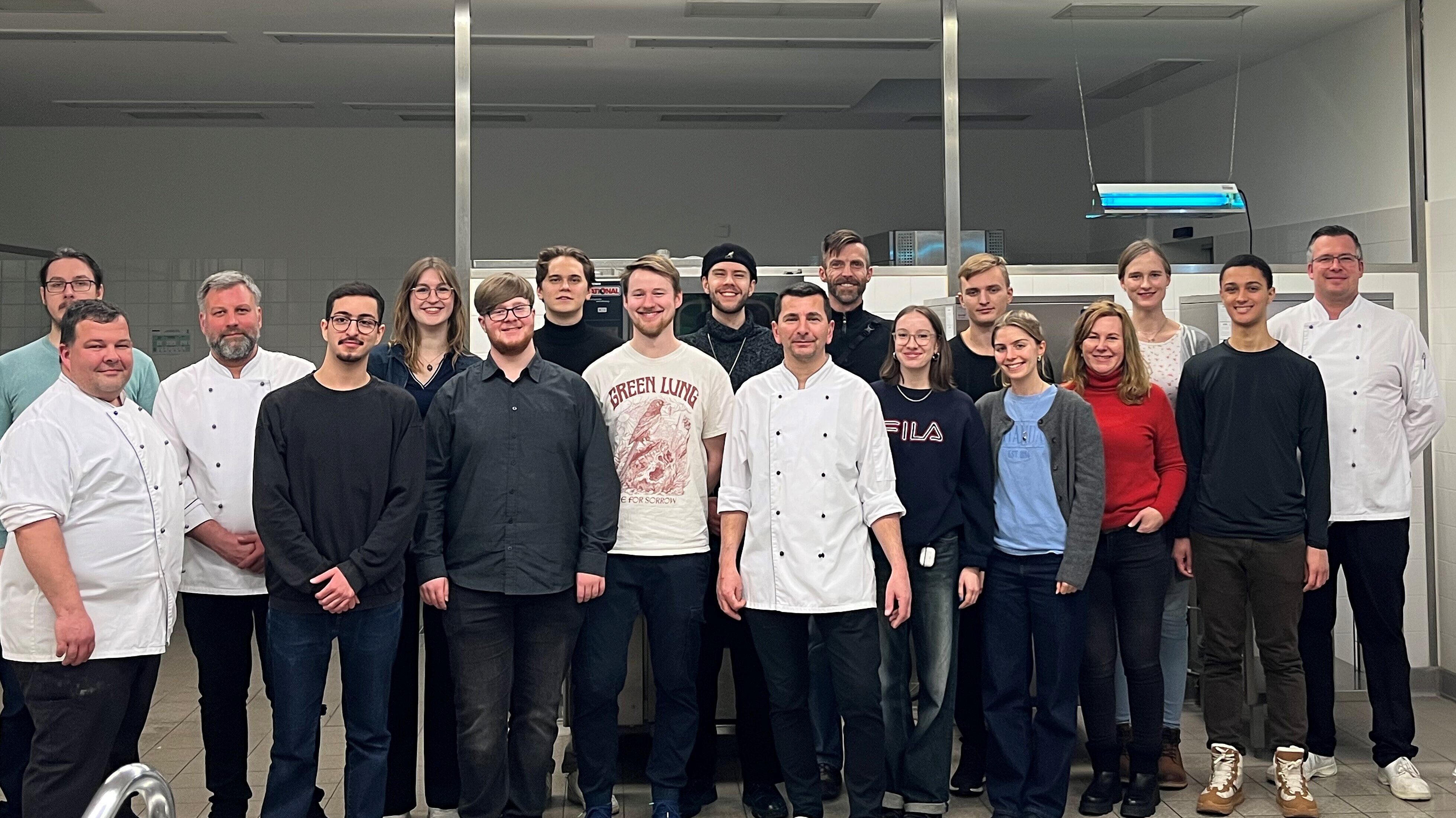 Eine Gruppe von Menschen steht in einer Küche und lächelt in die Kamera. Die meisten tragen Kochkleidung, was darauf hindeutet, dass es sich um Köche oder Küchenpersonal handelt. Die Atmosphäre wirkt freundlich und professionell.