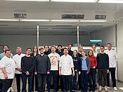 Eine Gruppe von Menschen steht in einer Küche und lächelt in die Kamera. Die meisten tragen Kochkleidung, was darauf hindeutet, dass es sich um Köche oder Küchenpersonal handelt. Die Atmosphäre wirkt freundlich und professionell.