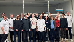 Eine Gruppe von Menschen steht in einer Küche und lächelt in die Kamera. Die meisten tragen Kochkleidung, was darauf hindeutet, dass es sich um Köche oder Küchenpersonal handelt. Die Atmosphäre wirkt freundlich und professionell.