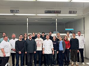 Eine Gruppe von Menschen steht in einer Küche und lächelt in die Kamera. Die meisten tragen Kochkleidung, was darauf hindeutet, dass es sich um Köche oder Küchenpersonal handelt. Die Atmosphäre wirkt freundlich und professionell.