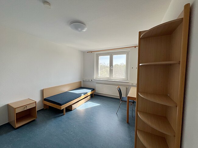 Einfaches, helles Zimmer mit einem schmalen Bett mit blauer Matratze, einem kleinen Tisch mit Stuhl daneben und einem Regal mit mehreren Fächern. Ein Fenster mit Vorhang lässt Tageslicht herein.