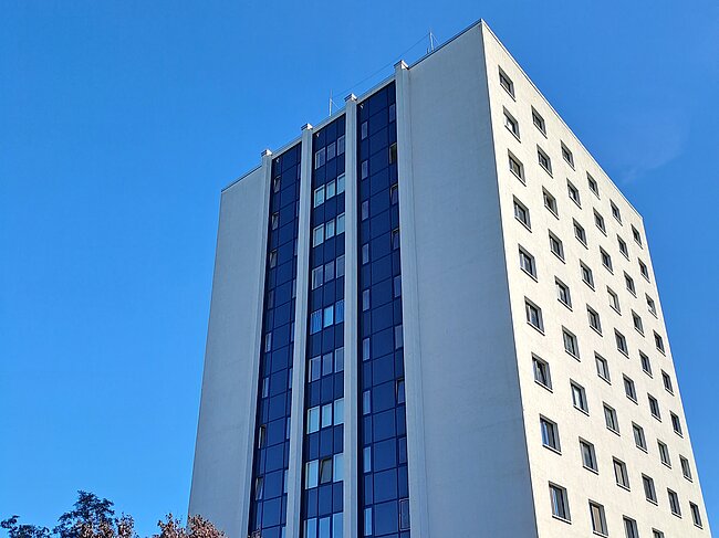 Ein modernes, mehrstöckiges Gebäude mit weißer Fassade und großen Glasflächen. Vor dem Gebäude befinden sich Fahrräder und ein Baum. Der Himmel ist klar und blau.