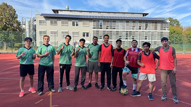 Gruppe von elf Personen auf einem Sportplatz, die fröhlich posieren. Sieben tragen grüne Trikots, vier rote. Im Vordergrund liegt ein Fußball.
