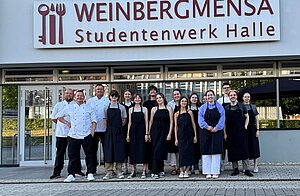 Ein Gruppenfoto vor dem Eingang der "Weinbergmensa Studentenwerk Halle". Die Gruppe besteht aus Mitarbeitern in Koch- und Servicekleidung. Sie stehen vor dem Gebäude und lächeln in die Kamera.