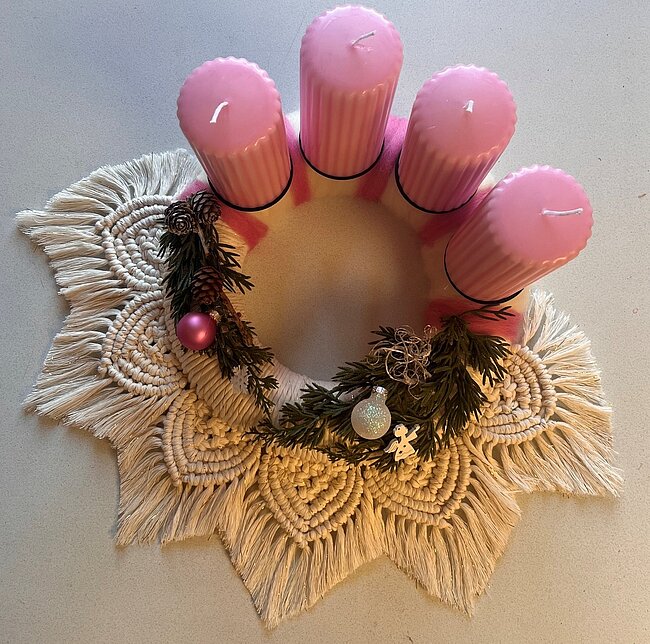 Dekorierter Adventskranz aus geflochtenen, hellen und weißen Makramee-Bändern mit drei rosa Kerzen in der Mitte, Tannenzweigen, einer kleinen Kugel und einem Stern als Schmuck.