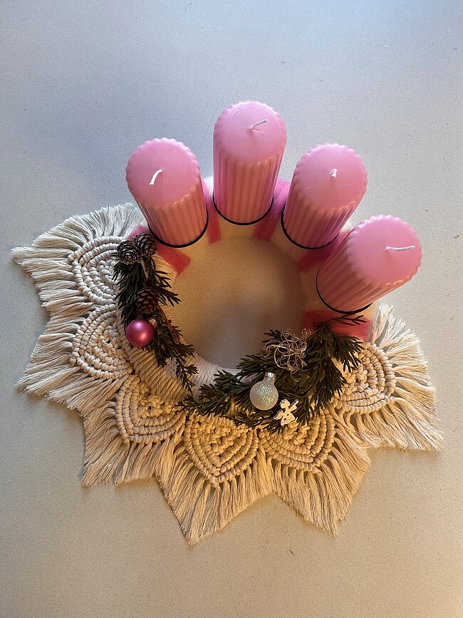 Dekorierter Adventskranz aus geflochtenen, hellen und weißen Makramee-Bändern mit drei rosa Kerzen in der Mitte, Tannenzweigen, einer kleinen Kugel und einem Stern als Schmuck.