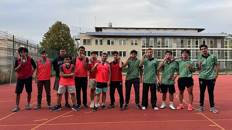 Gruppe von 14 Personen, darunter ein Rollstuhlfahrer in der Mitte, steht auf einem Sportplatz. Sie tragen Sportkleidung – einige in roten, andere in grünen Trikots – und posieren mit Siegergesten. Im Hintergrund ein Schulgebäude mit Solarpanels.
