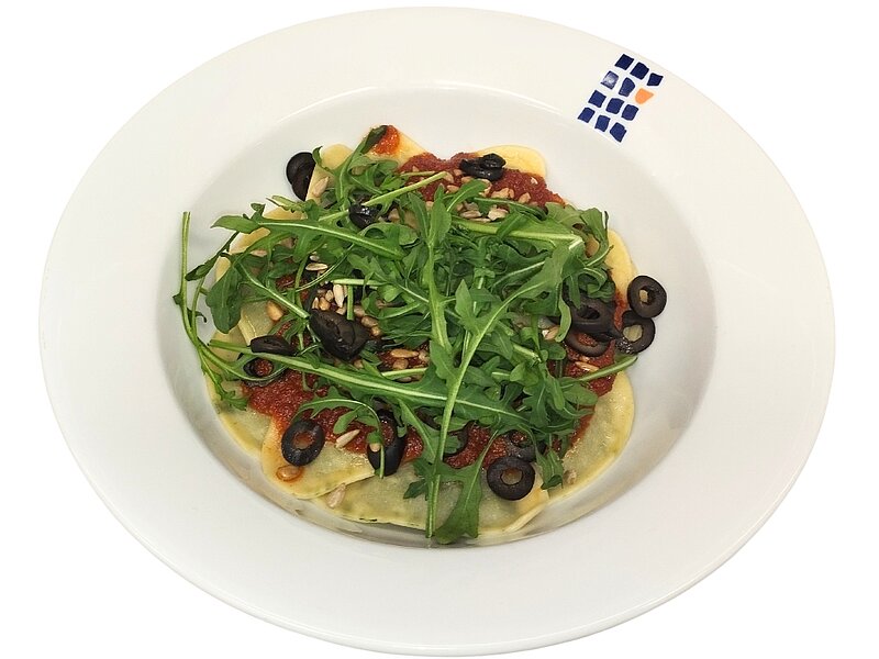 Ein Teller mit einer kleinen Pizza oder Focaccia belegt mit Rucola, schwarzen Oliven, getrockneten Tomaten und Pinienkernen.