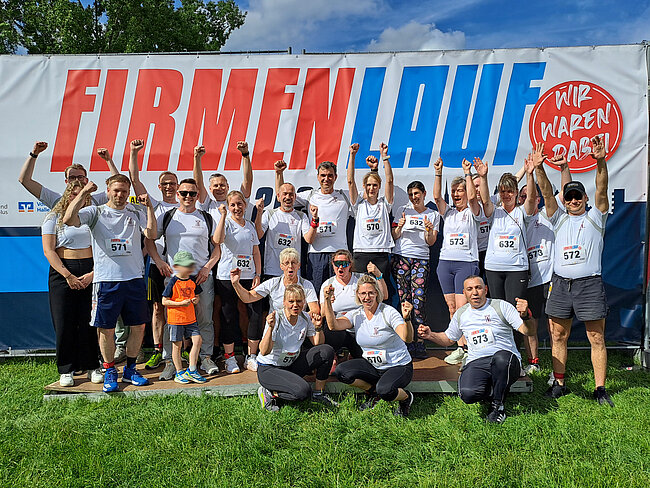 Gruppe von 18 fröhlichen Läuferinnen und Läufern mit Startnummern vor einem Banner der „Firmenlauf“-Veranstaltung, die mit beiden Händen nach oben gestreckt jubeln.