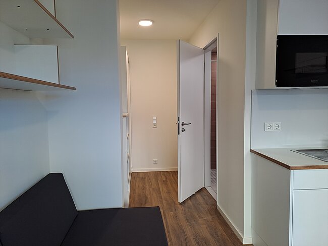 Moderne, helle Wohnküche mit offener Tür zum Badezimmer. Links ein dunkles Sofa, rechts eine Küchenzeile mit Arbeitsplatte, Herd und Mikrowelle. Boden in Holzoptik, weiße Wände.