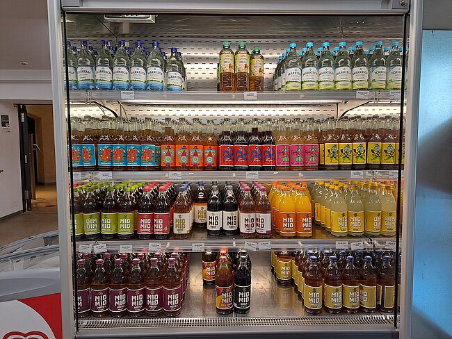 Offene Kühltheke mit fünf Regalen voller Getränkeflaschen: oben Wasser und Säfte, darunter verschiedene Limonaden, Energydrinks und Softdrinks in bunten Flaschen, unten dunkle Brause- und Erfrischungsgetränke mit Etiketten in verschiedenen Sprachen.