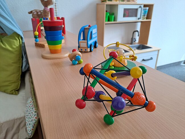 Bunte Holzspielzeug-Konstruktion aus Stäben und Kugeln in verschiedenen Farben wie Rot, Blau, Grün, Gelb und Orange auf einem Tisch. Im Hintergrund weitere Spielzeuge wie ein Stapelring und ein Spielzeugbus.