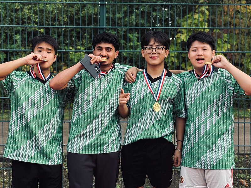 Vier junge Männer in grün-weiß gestreiften Sportshirts und Sportkleidung stehen auf einem Tennisplatz, zeigen Siegergesten und tragen jeweils eine Medaille um den Hals. Sie wirken stolz und erfolgreich.