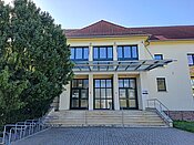 Ein Gebäude mit gelber Fassade, rotem Dach und mehreren großen Fenstern. Der Eingang besteht aus einer doppelten Glastür mit einem überdachten Vordach. Vor dem Gebäude befindet sich ein Baum und eine kleine Treppe mit einem Handlauf. Der Boden ist gepflastert.