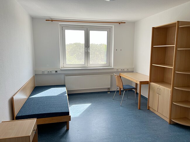 Ein helles, schlichtes Zimmer mit einem Einzelbett links, einem kleinen Schreibtisch mit Stuhl und einem großen Fenster mit Blick nach draußen. Rechts steht ein Regal mit Schubladen und offenen Fächern.