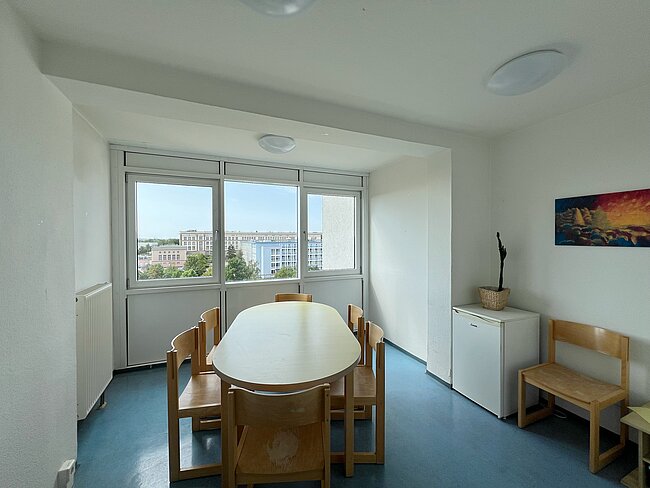 Helles Wohnzimmer mit großer Fensterfront, Blick auf Wohngebäude und Himmel. Zentral steht ein runder Esstisch mit vier Stühlen aus hellem Holz. Rechts daneben ein kleiner weißer Kühlschrank und ein Holzbank-Sitzplatz.