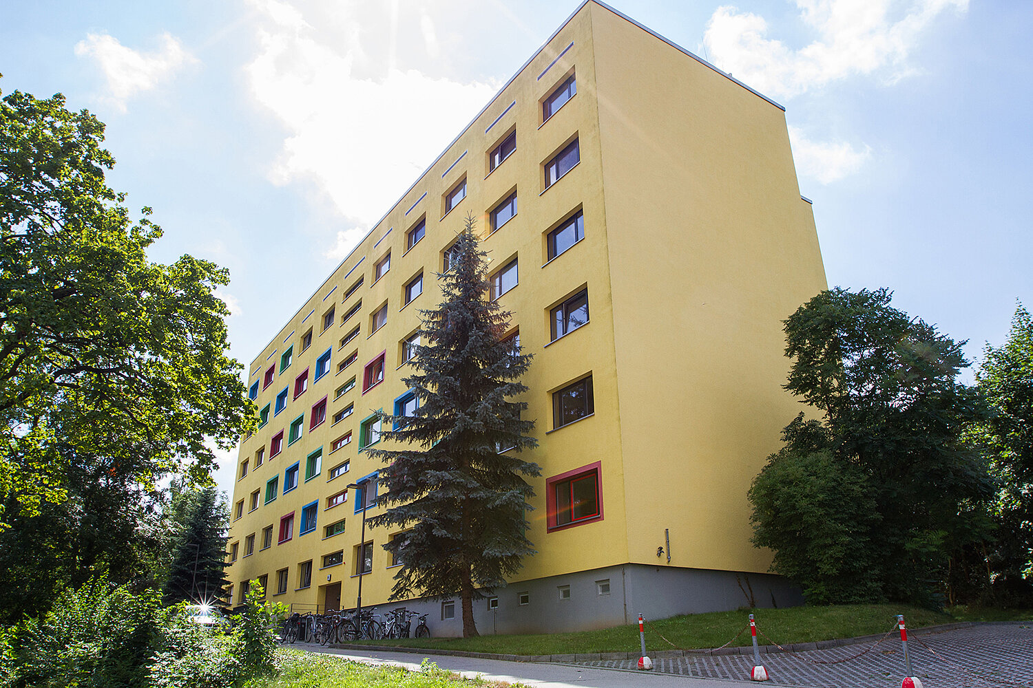 Ein mehrstöckiges, gelbes Wohngebäude mit vielen Fenstern, umgeben von Bäumen und Sträuchern. Vor dem Gebäude befindet sich ein gepflasterter Bereich mit einigen Verkehrshütchen. Der Himmel ist teilweise bewölkt.
