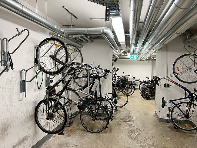 Ein unterirdischer Fahrradabstellraum mit mehreren Fahrrädern, die an Halterungen und an der Wand befestigt sind. Die Räder sind in verschiedenen Größen und Ausführungen, teilweise mit Gepäckträgern und Schutzblechen.