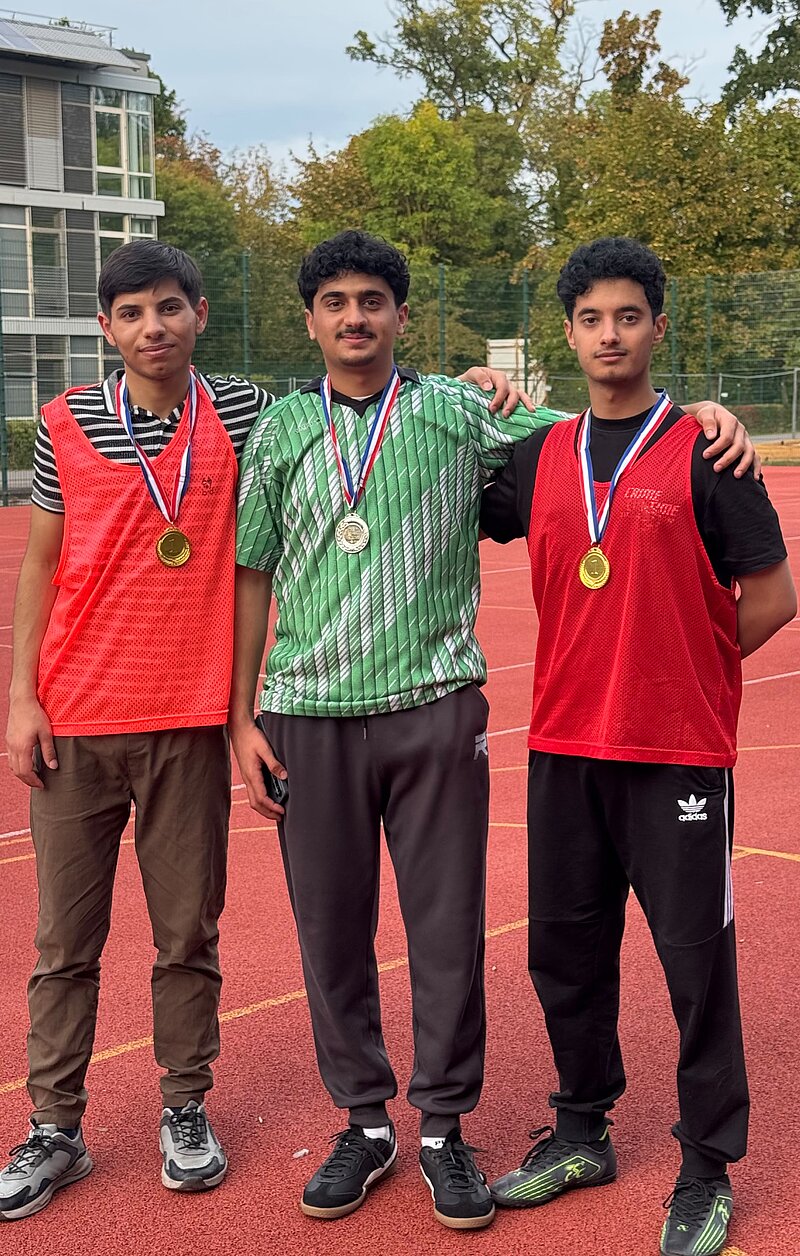 Drei junge Männer stehen auf einer Tartanbahn, jeder mit einer goldenen Medaille um den Hals, lächelnd und stolz. Sie tragen Sportkleidung und posieren gemeinsam nach einem Wettkampf.