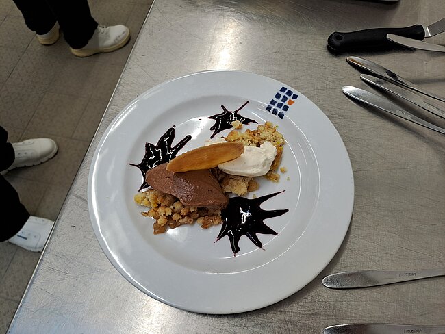 Ein Dessertteller mit einer Schicht aus knusprigem Haferflocken-Gericht, darüber eine Scheibe geröstetes Brot oder Biskuit mit Schokoladenüberzug, daneben eine Portion Vanilleeis und geschmackvoll mit Schokoladensauce in Sternform garniert.