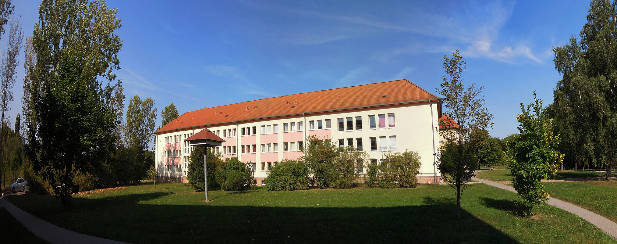 Ein großes, weißes Schulgebäude mit rotem Dach steht in einem grünen, parkähnlichen Gelände. Der Himmel ist blau mit einigen Wolken. Vor dem Gebäude befindet sich ein gepflegter Rasen mit Bäumen und Sträuchern.