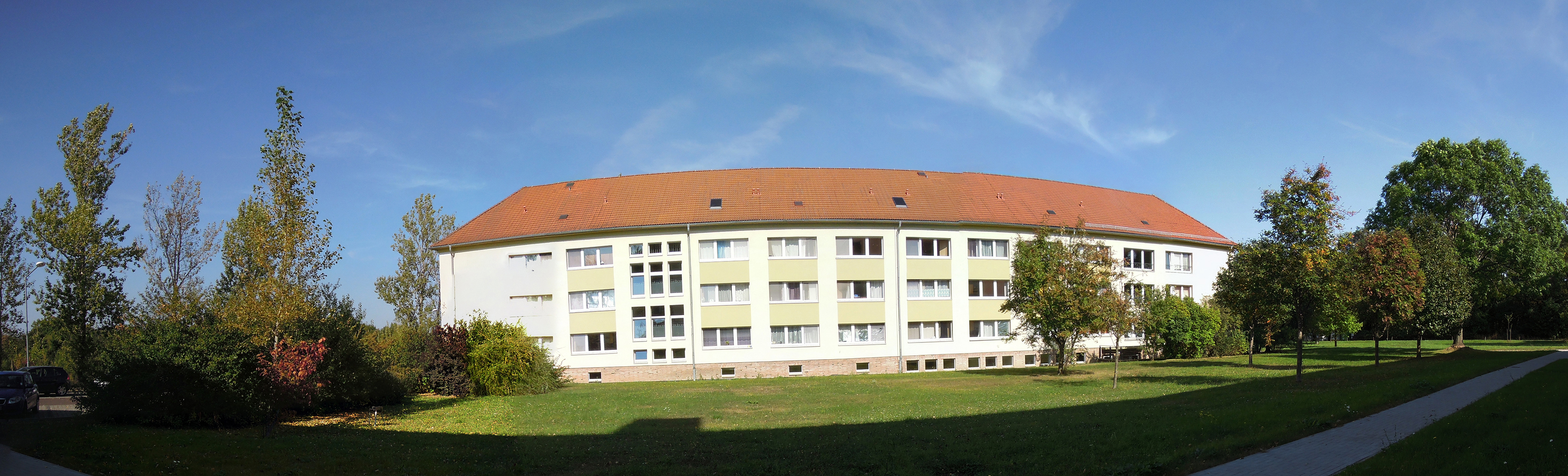Ein großes, mehrstöckiges Gebäude mit rotem Dach und vielen Fenstern. Es steht auf einem weitläufigen, grünen Rasen mit Bäumen und Sträuchern in der Umgebung. Der Himmel ist blau mit einigen Wolken.
