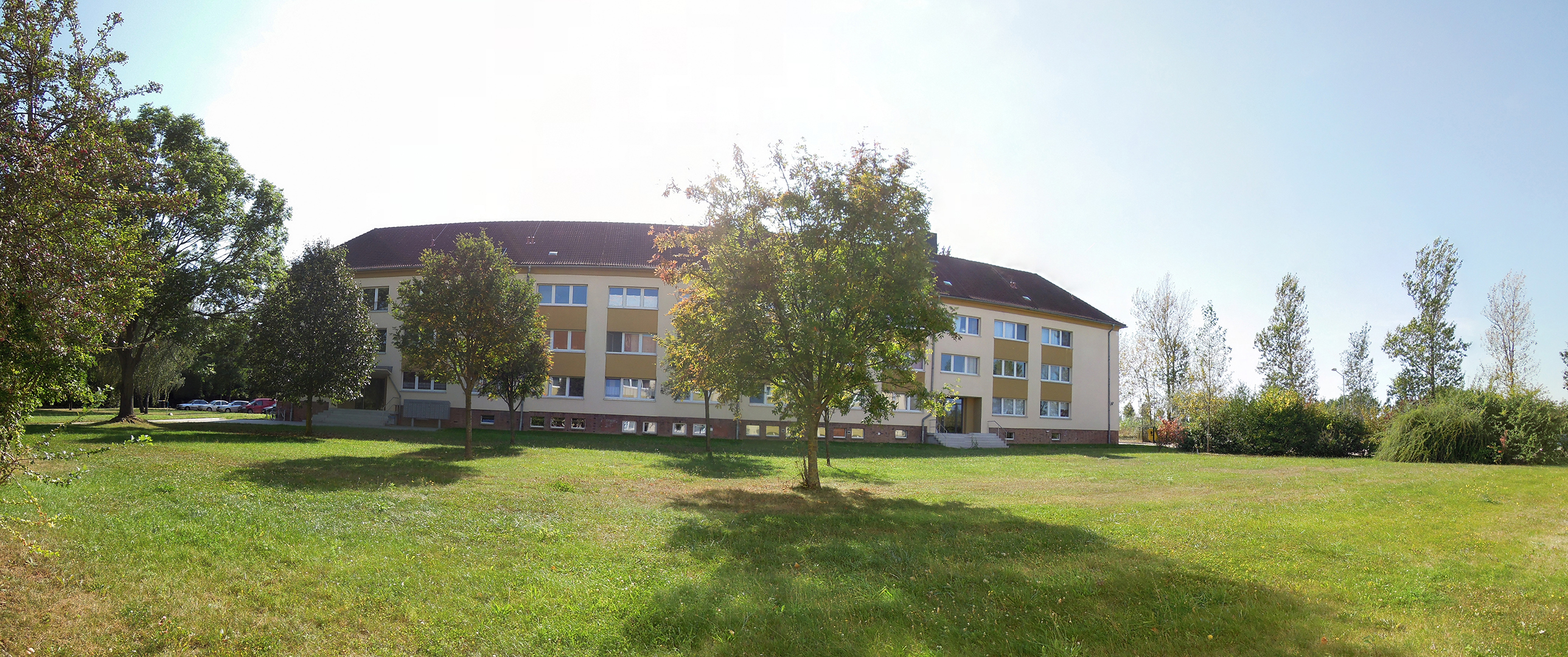 Ein großes, weißes Gebäude mit mehreren Fenstern steht auf einer Wiese. Im Vordergrund befinden sich Bäume und ein offenes Grasfeld. Der Himmel ist klar und sonnig.