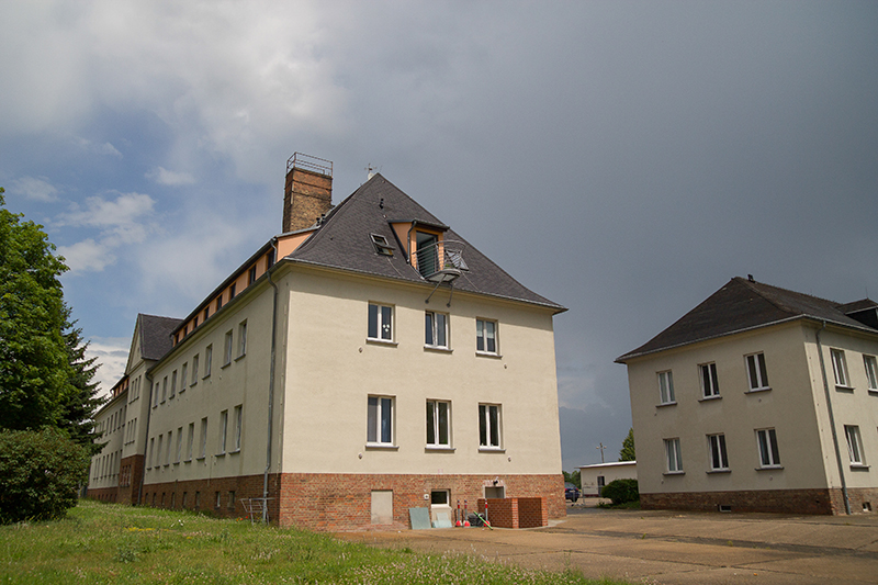 Zwei mehrstöckige Gebäude mit hellen Fassaden und dunklen Dächern stehen auf einem gepflegten Gelände. Das linke Gebäude hat einen Schornstein und mehrere Fenster, während das rechte Gebäude etwas kleiner ist. Der Himmel ist bewölkt, und im Vordergrund befindet sich ein grasbewachsener Bereich.
