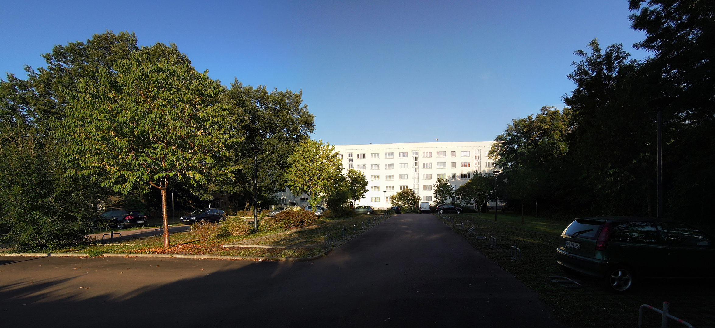 Ein Wohngebäude steht am Ende einer ruhigen Straße, umgeben von Bäumen und einigen geparkten Autos. Die Sonne scheint, und der Himmel ist klar. Die Straße ist asphaltiert und führt direkt zum Gebäude. Es ist ein friedliches Wohngebiet mit viel Grün.