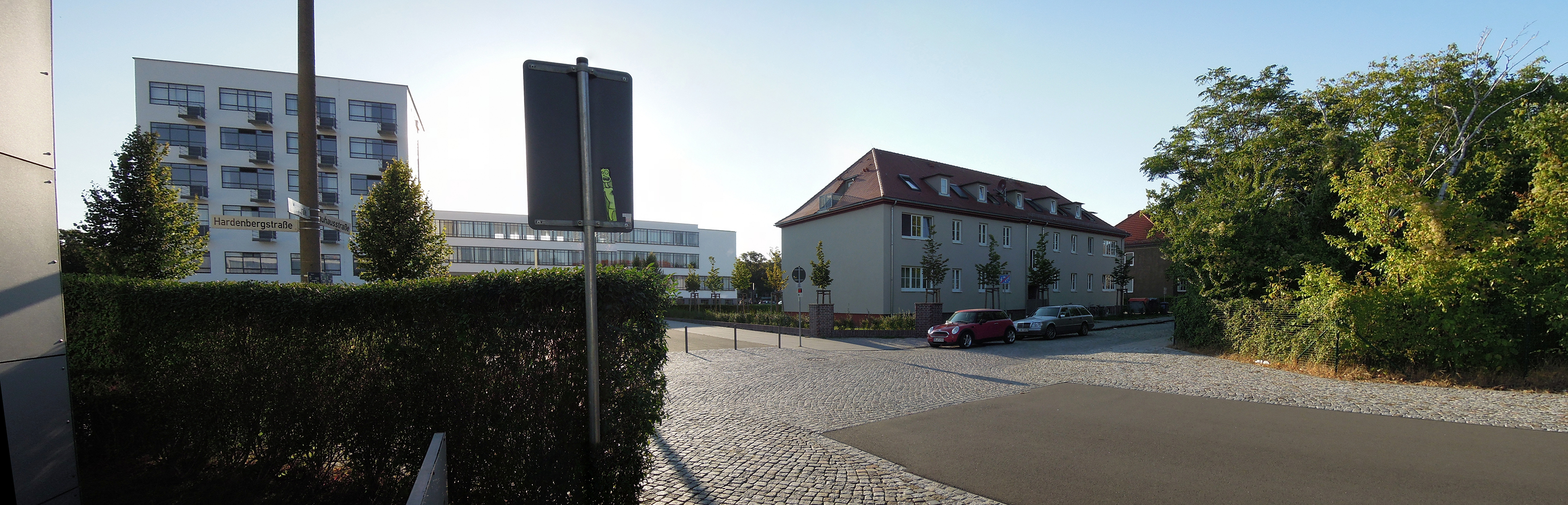 Ein Panorama einer Wohngegend mit einem Parkplatz vor einem Mehrfamilienhaus und einem Wohnblock im Hintergrund. Der Parkplatz ist von gepflegten Sträuchern und Bäumen umgeben. Der Himmel ist klar und blau.