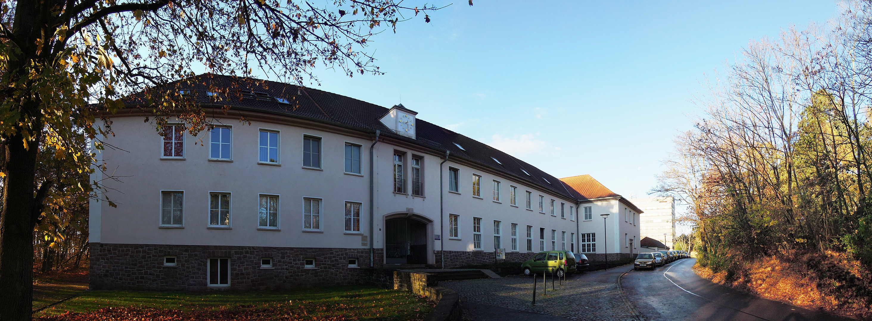 Ein großes, weißes Gebäude mit mehreren Stockwerken und vielen Fenstern steht an einer ruhigen Straße. Die Umgebung ist von Bäumen und Natur umgeben, und es ist ein klarer, sonniger Tag. Die Straße führt direkt zum Gebäude, das einen gepflegten Eindruck macht.