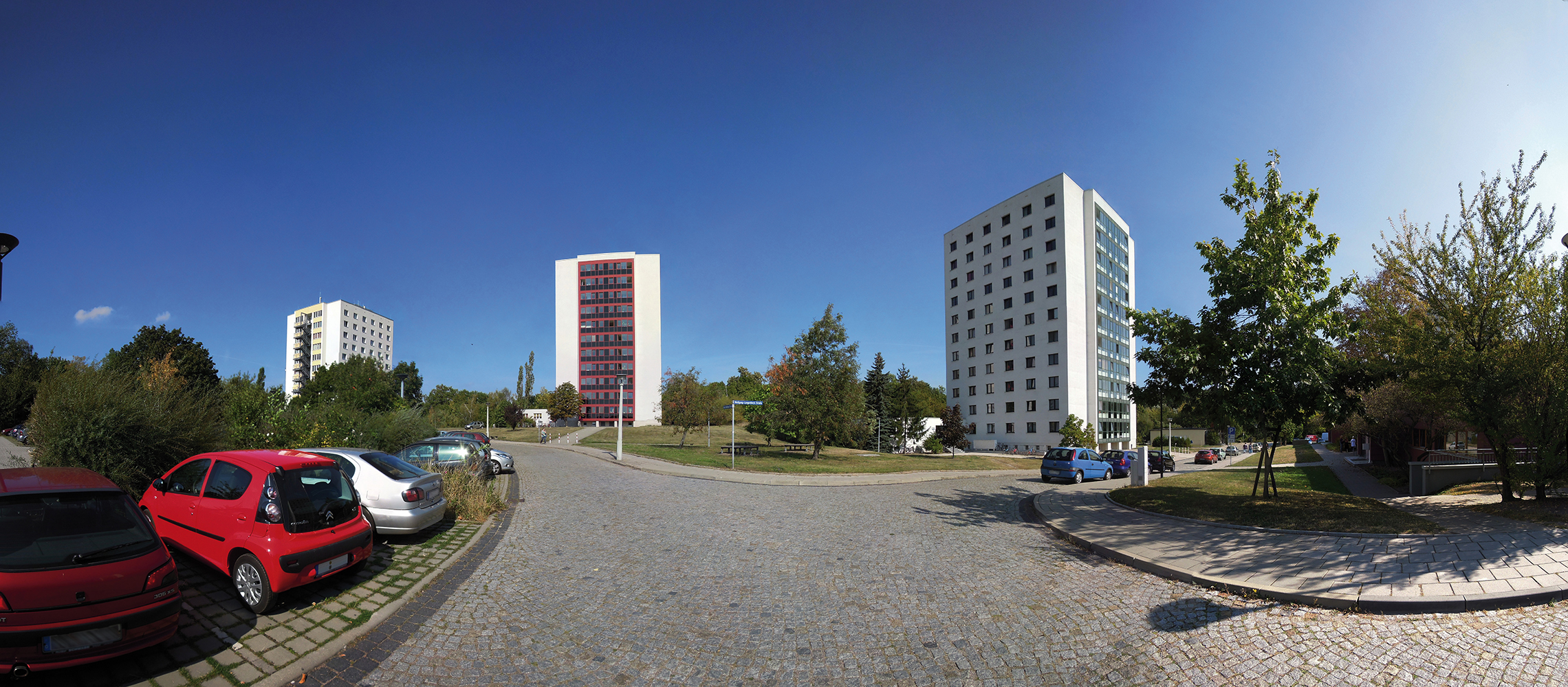 Ein Panoramabild einer Wohngegend mit mehreren Hochhäusern, geparkten Autos und einer gepflasterten Straße. Im Vordergrund sind Bäume und ein kleiner Parkbereich zu sehen. Der Himmel ist klar und blau.