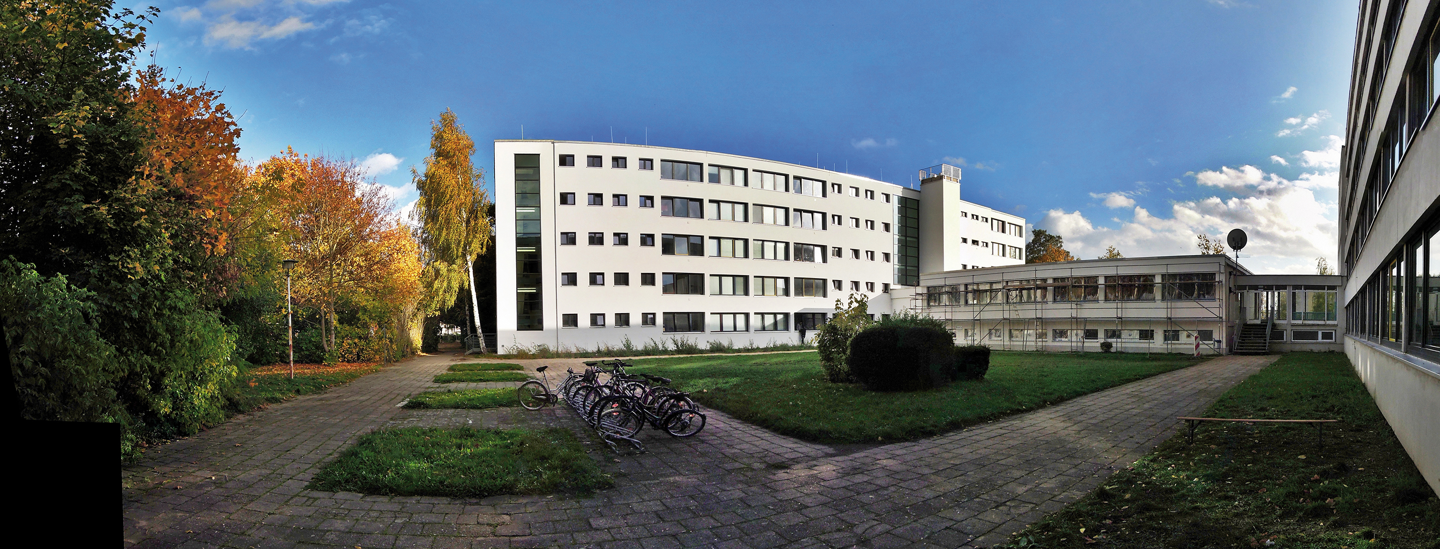 Ein Panoramabild eines Universitätsgeländes mit mehreren Gebäuden, einem Rasenbereich und einigen Fahrrädern. Im Hintergrund sind Bäume mit herbstlicher Färbung zu sehen. Der Himmel ist teilweise bewölkt.