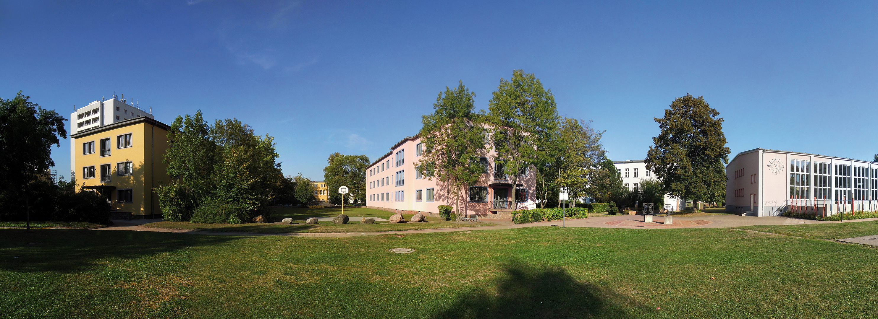 Ein Panoramabild eines Campus mit mehreren Gebäuden, grünen Bäumen und einem weitläufigen Rasenbereich. Die Gebäude sind modern und mehrstöckig, umgeben von viel Natur. Der Himmel ist klar und blau, was auf einen sonnigen Tag hindeutet.