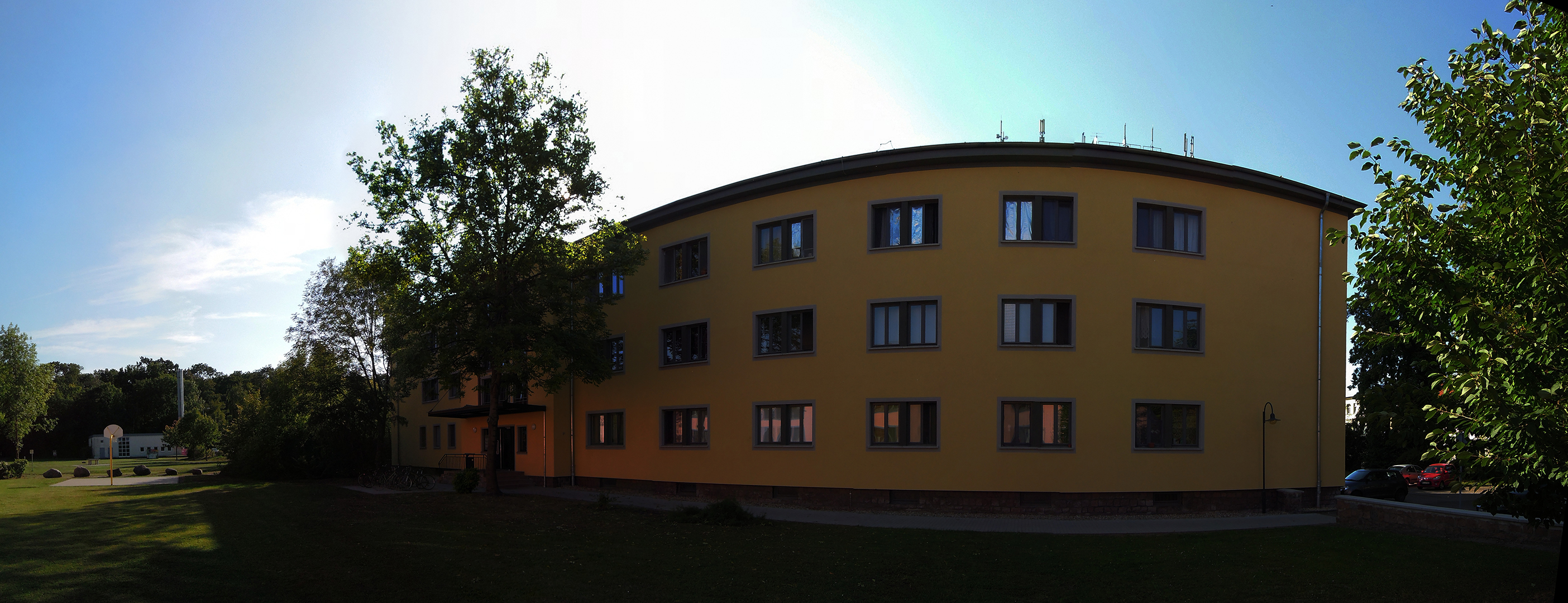 Ein mehrstöckiges, gelbes Gebäude mit vielen Fenstern steht vor einem klaren Himmel. Es ist von Bäumen und Grünflächen umgeben. Die Sonne scheint hell, und es ist ein ruhiger, sonniger Tag.